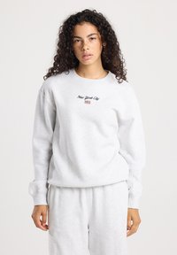 Joven con cabello rizado que lleva una sudadera gris claro y pantalones, la sudadera está bordada con "Nueva York" y una pequeña bandera americana.