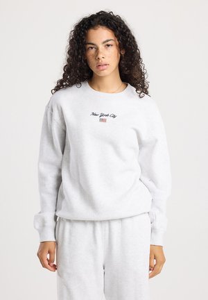 Jonge vrouw met krullend haar, gekleed in een lichtgrijze trui en broek. De trui is geborduurd met "New York City" en een kleine Amerikaanse vlag.