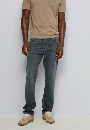 Homme portant un jean bleu délavé, un t-shirt beige et des baskets beiges, debout devant un fond uni clair.