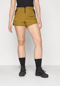 Olive green shorts with a drawstring waist, black side panels, and an angled hem. Worn with black hiking shoes and knee-length socks.