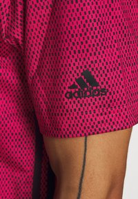 Růžové sportovní tričko Adidas s geometrickým černým vzorem a černým logem na spodním lemu. Materiál se zdá být lehký a texturovaný.