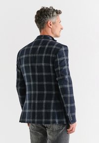 Granatowa marynarka w kratę z miękkiego materiału, dopasowany krój, pojedyncze rozcięcie, mankiety z trzema guzikami i wyraźnie zarysowane ramiona. Noszona na szarych dżinsach.