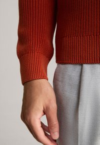 Polsino di maglione rosso a coste con dettaglio di lavorazione a maglia, indossato con tessuto grigio chiaro, che mostra un design aderente e rifiniture pulite.