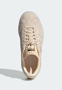 Beige suede sneakers feature a textured upper, woven laces, gold logo on the tongue, and white rubber sole with three stripes.