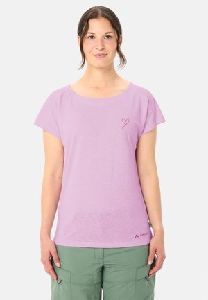 Vrouw die een lichtpaars T-shirt met korte mouwen en een klein hartje draagt, en groene cargobroek, staand tegen een effen achtergrond.