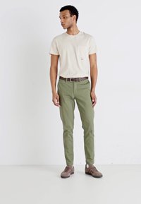 Camiseta de color beige claro con detalle de bolsillo, combinada con pantalones verde oliva de corte ajustado. Cinturón y zapatos marrones, contra un fondo neutro.