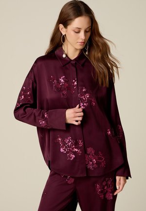 Frau mit langen braunen Haaren trägt eine dunkelviolette Satinbluse und Hose mit pinken floralen Pailletten-Details sowie große Creolen.