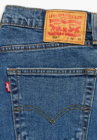 Jeans Levi's 514 en denim azul oscuro, con un parche trasero de cuero marrón, etiqueta roja en el bolsillo izquierdo y costuras amarillas en contraste.