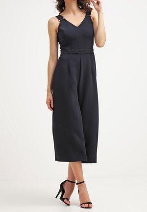 Femme portant une combinaison sans manches noire à jambes larges avec des bretelles à boucle et des sandales noires à talons hauts et brides à la cheville, sur un fond blanc.