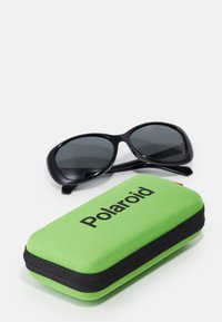 Juodi akiniai nuo saulės su tamsiais objektyvais greta tekstūruoto žalio kietos dėžutės, ant kurios matyti užrašas "Polaroid" ryškiomis juodomis raidėmis.