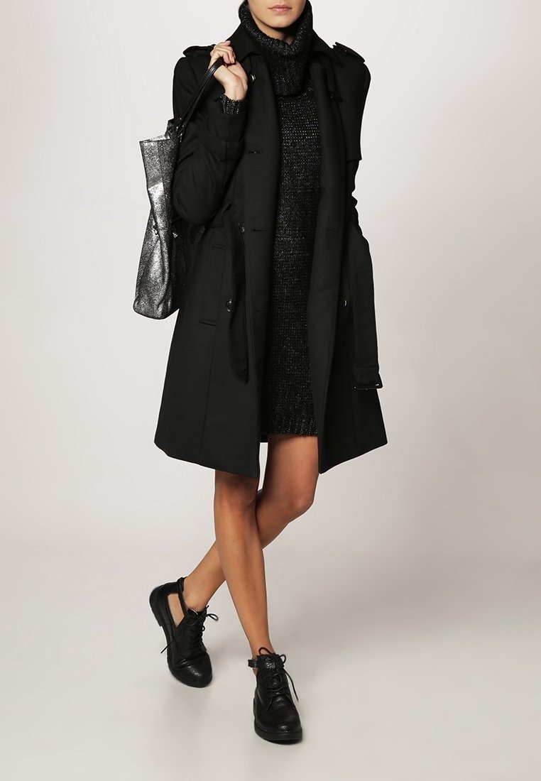 Femme en trench noir et robe pull, debout jambes croisées, tenant un sac métallique et portant des baskets noires devant un fond uni.