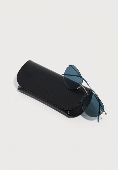 Étui à lunettes noir avec logo embossé et lunettes de soleil rondes à verres bleutés posées dessus, sur un fond clair uni.