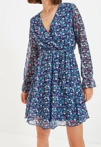 Kukkatulokset mekko kevyessä tummansinisessä kankaassa, jossa on pitkät hihat. Siinä on monivärisiä pieniä kukkia, vyötärönsitoja ja levenevä helma.