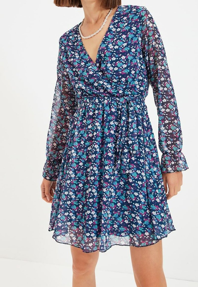 Kukkatulokset mekko kevyessä tummansinisessä kankaassa, jossa on pitkät hihat. Siinä on monivärisiä pieniä kukkia, vyötärönsitoja ja levenevä helma.