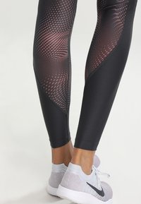 Nogi w czarnych legginsach w wzory i szarych butach do biegania Nike z białymi podeszwami, stojące na jasnym tle.