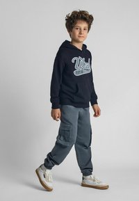 Haut à capuche noir avec logo blanc, pantalon cargo gris avec poches latérales, et baskets blanches avec semelles beige. Tenue simple et décontractée sur un garçon.