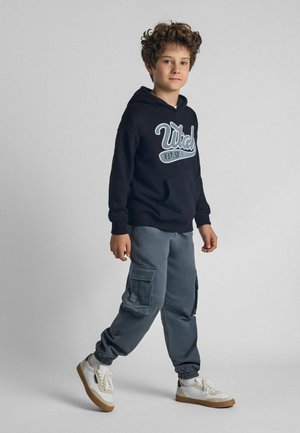 Haut à capuche noir avec logo blanc, pantalon cargo gris avec poches latérales, et baskets blanches avec semelles beige. Tenue simple et décontractée sur un garçon.