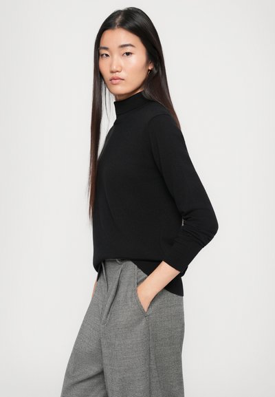 Schwarzer Rollkragenpullover mit langen Ärmeln, glatter Textur, kombiniert mit grauen Hahnentritthosen, die einen lässigen Schnitt und seitliche Taschen aufweisen.