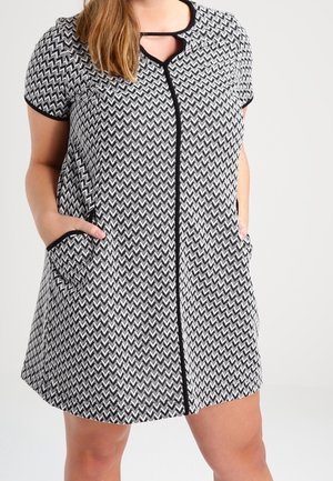 Kobieta ubrana w sukienkę na krótki rękaw w czarno-biały zygzakowaty wzór z kieszeniami i czarnym wykończeniem, stojąca z rękami w kieszeniach.