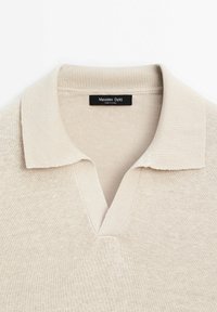 Beige neulepolo, jossa on V-kaula-aukko, ribattu kaulus ja kiinnitetty etiketti. Kangas näyttää pehmeältä, sillä on sileä rakenne ja vähäiset muotoiluyksityiskohdat.