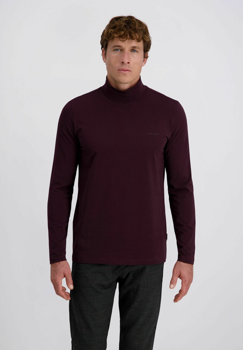 Mann mit lockigem Haar, der einen dunkelvioletten Langarmrollkragenpullover und eine dunkelgraue karierte Hose trägt, steht vor einem einfarbigen Hintergrund.