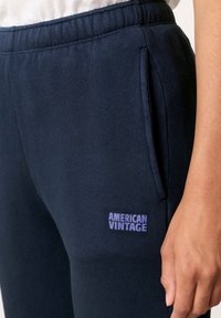 Tmavomodré tepláky vyrobené z mäkkého materiálu, s bočnými vreckami a malým fialovým logom "AMERICAN VINTAGE" blízko pásu.