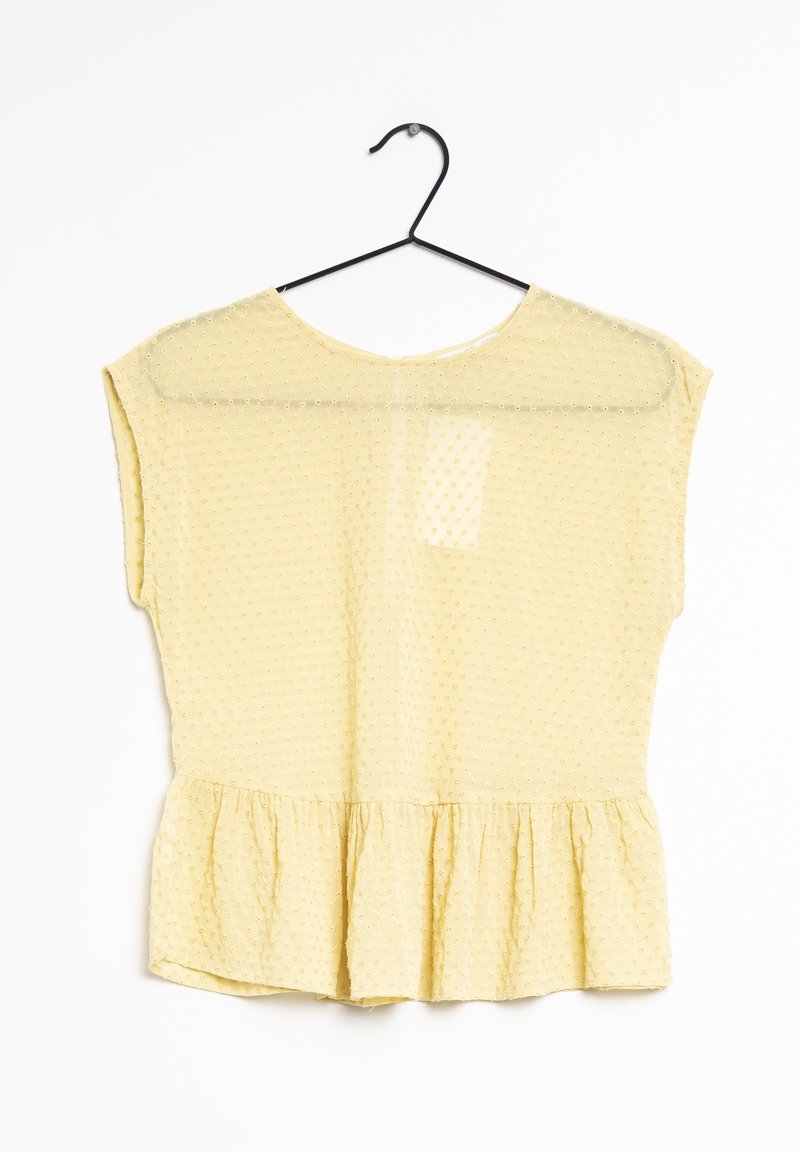 Blouse jaune à manches courtes, motif texturé, encolure ronde et ourlet volanté. Accrochée à un crochet noir sur un fond blanc.