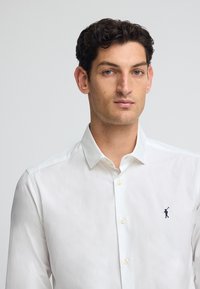 Vit långärmad skjorta med knapp-down krage, med en marinblå broderad logotyp på bröstet och plastknappar. Slät tygtextur.