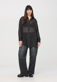 Camicia trasparente nera con bottoni e maniche lunghe, indossata sopra un indumento intimo nero, abbinata a jeans cargo neri larghi e stivali neri chunky.