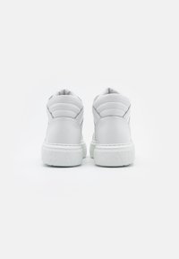 Bílé kožené vysoké tenisky s polstrovanými patami a silnými gumovými podešvami s vyraženými texturovanými písmeny, zobrazené zezadu na bílém pozadí.