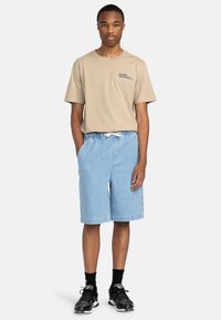 Beige T-Shirt mit kurzen Ärmeln und Logo, kombiniert mit hellblauen Denim-Shorts. Schwarze Sneakers und Socken runden den Look ab. Lässiger Stil.