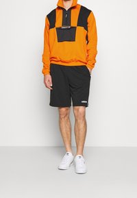 Felpa con zip arancione e nera con tasca frontale, abbinata a pantaloncini neri. Scarpe da ginnastica bianche con dettagli di branding discreti.