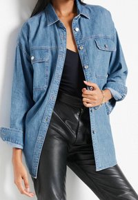 Chemise en denim bleu clair avec fermeture à boutons, deux poches avant et manches retroussées, associée à un pantalon noir en simili cuir.