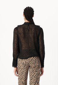 Zwarte transparante blouse met stippenstructuur, gerimpelde kraag en lange mouwen. Gecombineerd met tan en zwarte zebra-print broek.
