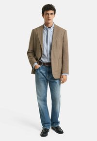 Braun karierter Blazer über einem blau-weißen gestreiften Hemd, kombiniert mit hellblauen gerade geschnittenen Jeans und schwarzen Schuhen. Stehende Haltung.