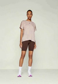 Nő bézs rövidujjú felsőben, sötétbarna kerékpáros rövidnadrágban, fehér zokniban és lila talpú fehér sportcipőben áll egy világos háttér előtt.