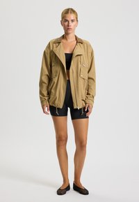Khakifarbene oversized Jacke mit Kragen, Fronttaschen und Reißverschluss. Über einem schwarzen Crop-Top und schwarzen Shorts getragen. Strukturierter Stoff.