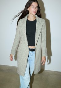 Siv paličasti plašč z usklajenimi reverji, dolgimi rokavi in stranskimi žepi, nosi se čez črno majico in svetlo modre kavbojke. Gladka tekstura.