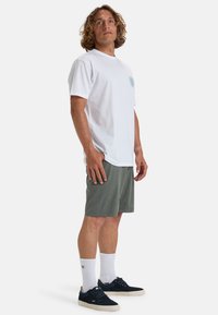 T-shirt bianco in cotone con un logo circolare color teal sul petto, abbinato a pantaloni corti verdi, calzini bianchi e sneakers blu navy.