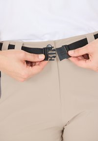 Beige bukser med et sort justerbart bælte, der har en plastikspænde og lukningsmekanisme, båret i taljen, hænder der sikrer spænde.