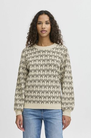 Pull beige avec un motif de nœuds répété en noir, encolure ronde et poignets côtelés. Associé à un jean bleu clair.