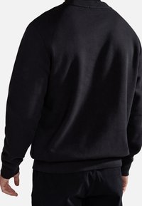 Homme portant un sweat-shirt noir uni et un pantalon noir, vu de dos avec les bras détendus le long du corps.