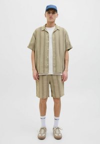 Chemise beige légère, rayée, à manches courtes avec un bouton sur le devant, associée à un short assorti. Portée avec des chaussettes blanches et bleues, et des baskets beiges.
