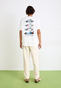 White t-shirt featuring three illustrated cars on the back, with the text "THE ULTIMATE DRIVING MACHINE" below. Casual fit, paired with cream pants.