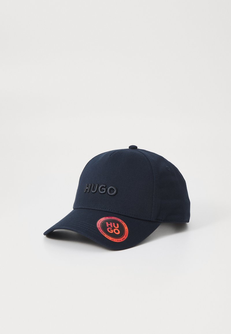 Mørkeblå baseballcaps med brodert "HUGO" logo, buet skygge og rød sirkulær tag med "HUGO" tekst. Stoffet har en jevn tekstur.