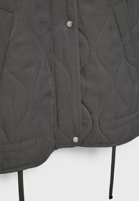 Donkergrijze gewatteerde stof met golvende stiknaden, zilveren drukknoopsluitingen, twee voorzakken en zwarte ophangstraps zichtbaar aan de onderkant.