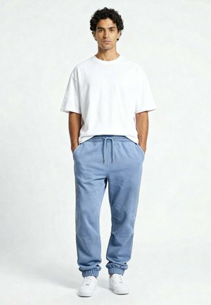 Ein Mann mit lockigem Haar steht mit den Händen in den Taschen und trägt ein weißes übergroßes T-Shirt, blaue Jogginghosen und weiße Turnschuhe vor einfarbigem Hintergrund.