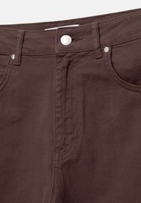 Gros plan d'un pantalon en denim marron montrant la taille, la fermeture par bouton, les poches avant et les passants de ceinture.