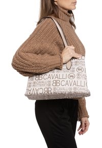 Sac à main beige avec un imprimé "CAVALLI" en marron sur l'ensemble. Le sac a une forme structurée avec une finition lisse et deux poignées.