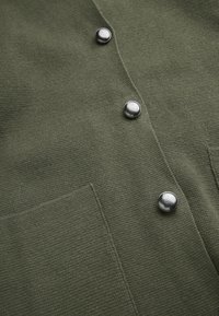 Olivgrön cardigan med en texturerad tyg, silverfärgade metallknappar och en platt fickdetalj på vänster sida.